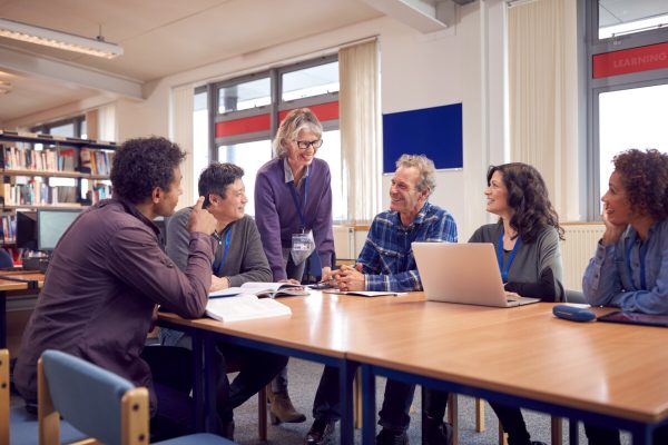 Teacher With Group Of Mature Adult Students In Class Sit Around Table And Work In College Library